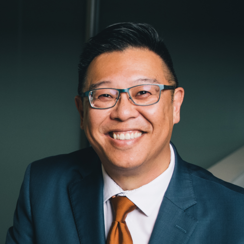 Man with glasses in suit and burnt orange tie smiling