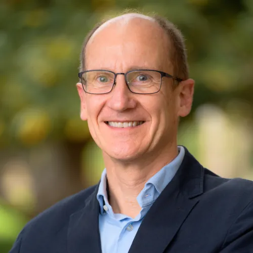 Man smiling. He is wearing glasses and a light blue shirt and blazer. 