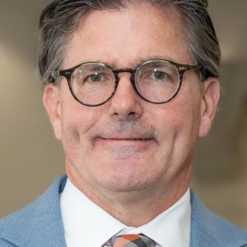 Picture of a man with round glasses and wearing a light blue suit and colorful tie smiling. 