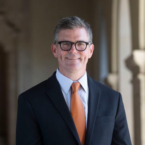 Man smiling at camera wearing a suit, light blue shirt, and burnt orange tie. a white shirt. 