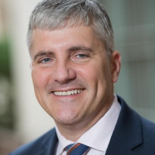 Man in dark blue suit, blue and burnt orange tie, and light collared shirt smiling. 
