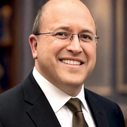 A man in a black suit, white shirt, and brown tie wearing glasses smiling.
